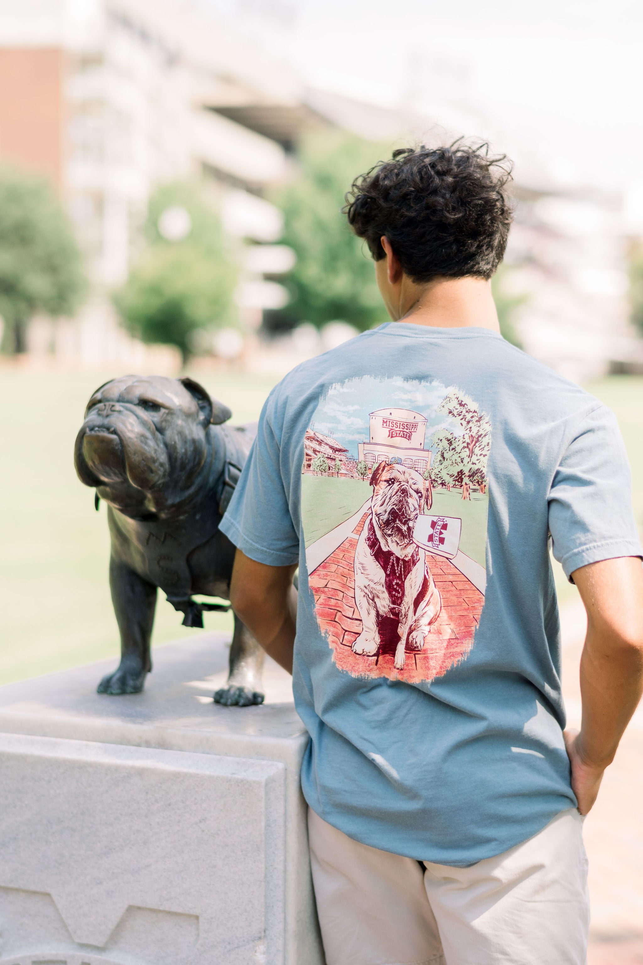 Mississippi State Dusty Blue T-Shirt with an image of Bully the bulldog holding a cowbell in its mouth on the brick walk into campus