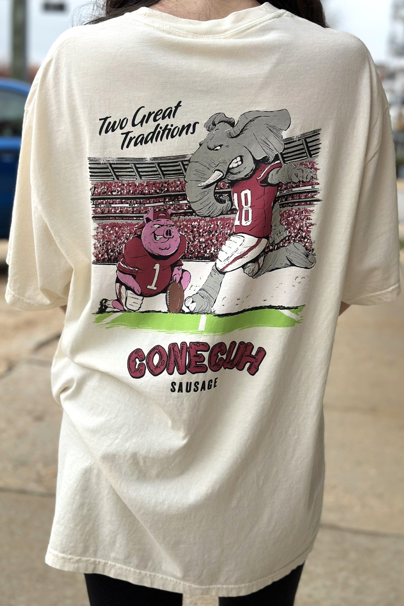 University of Alabama Conecuh Traditions Ivory Comfort Colors T-Shirt with Black text "Two Great Traditions" above an illustration of a pig holding a football for Big Al to kick 