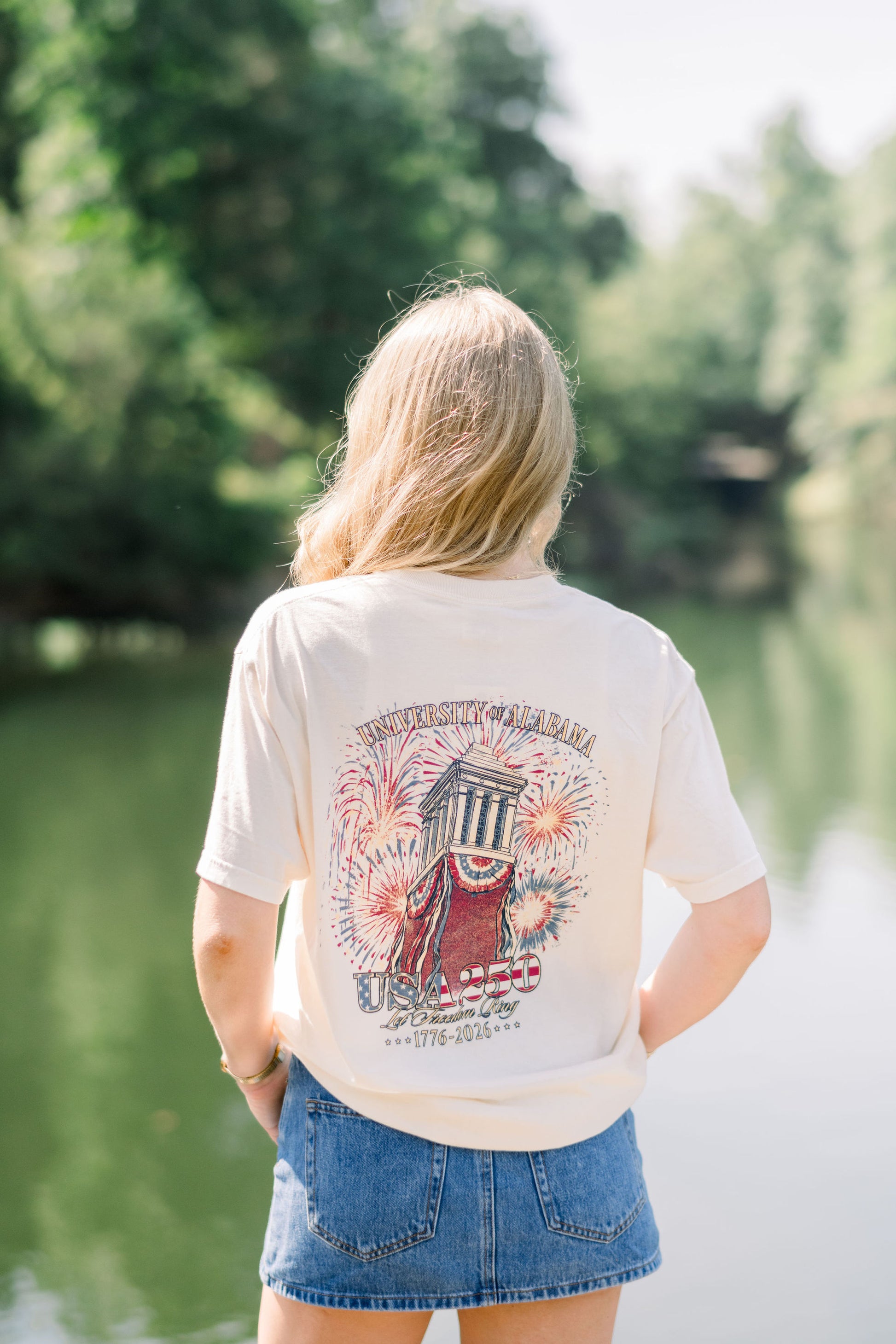 University of Alabama Ivory T-Shirt with an image of Denny Chimes decorated in Americana banners and fireworks exploding behind it with text “USA 250”