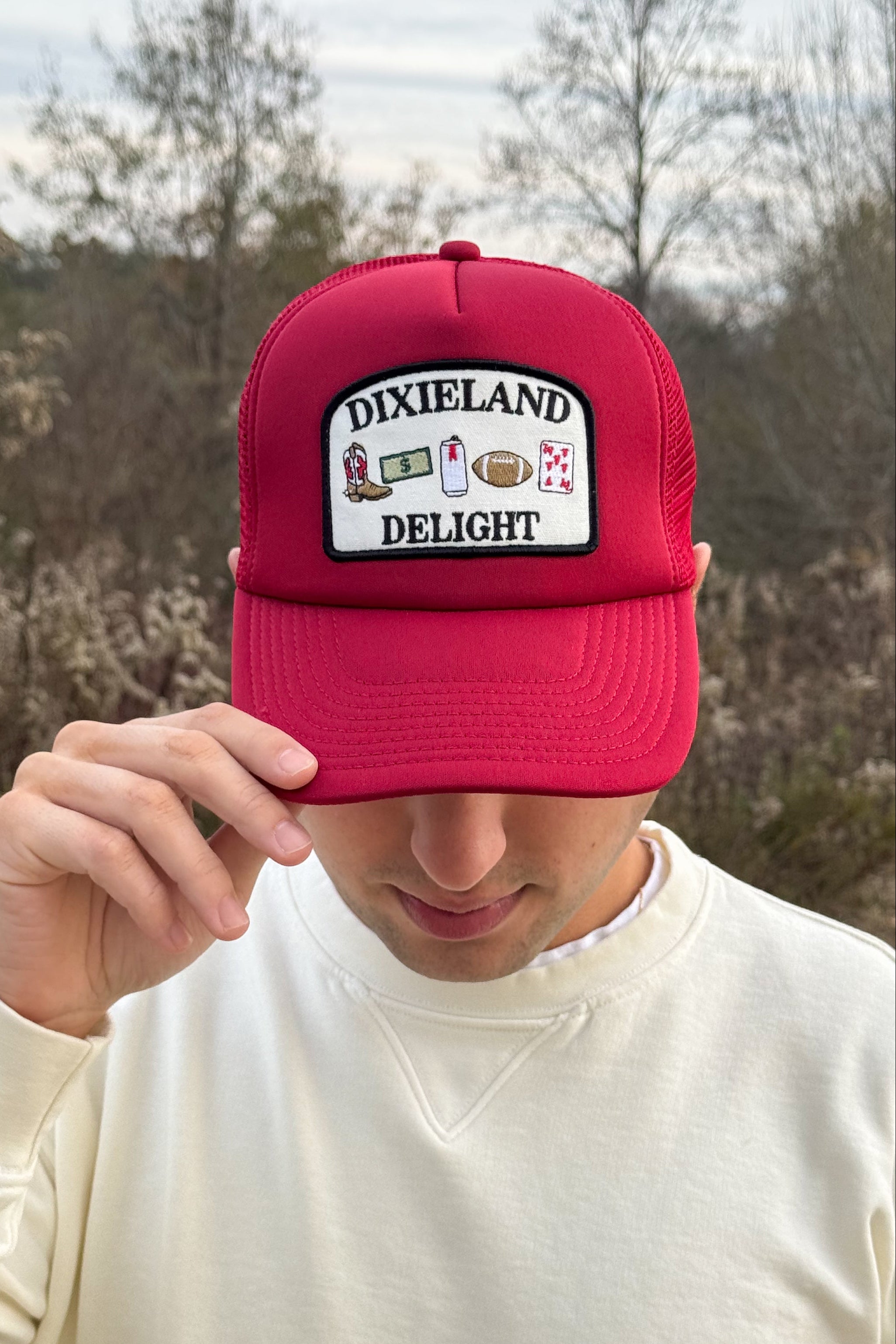 University of Alabama Red Trucker Hat with a patch featuring text “Dixieland” above and “Delight” below drawings of a cowboy boot, money, drink can, football, and playing card