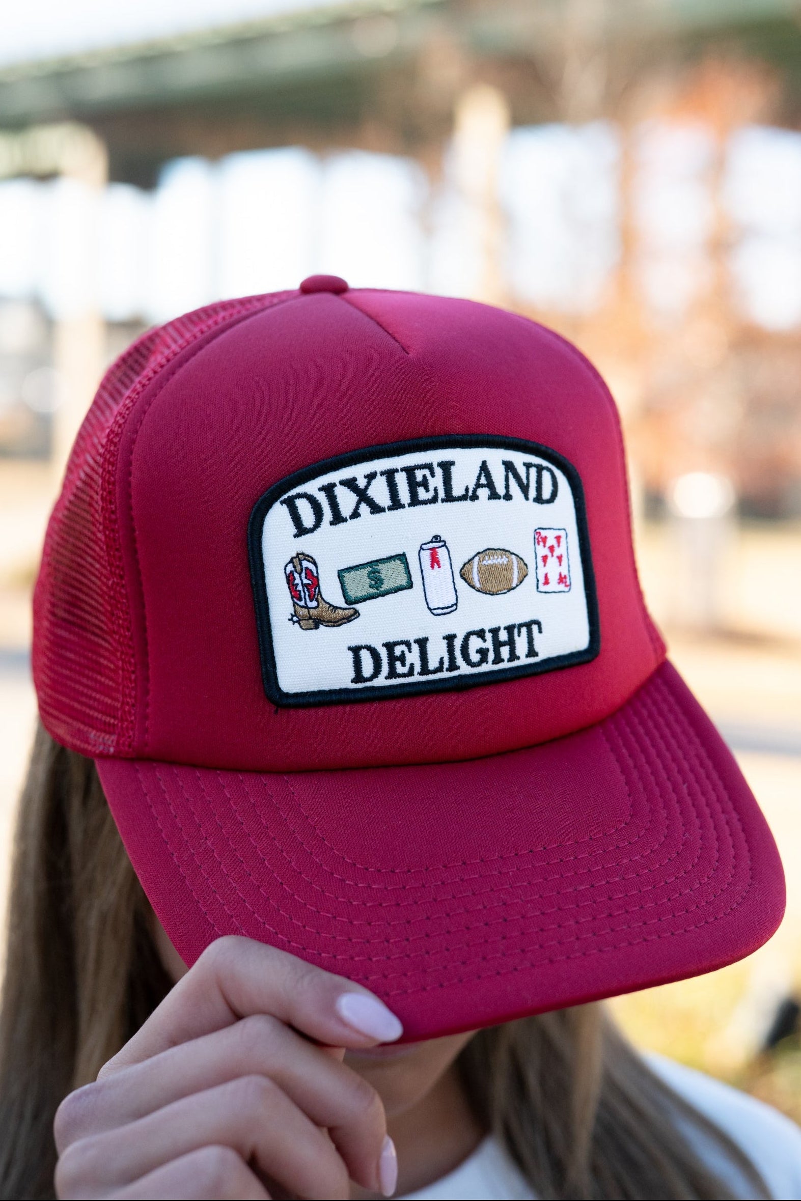 University of Alabama Red Trucker Hat with a patch featuring text “Dixieland” above and “Delight” below drawings of a cowboy boot, money, drink can, football, and playing card