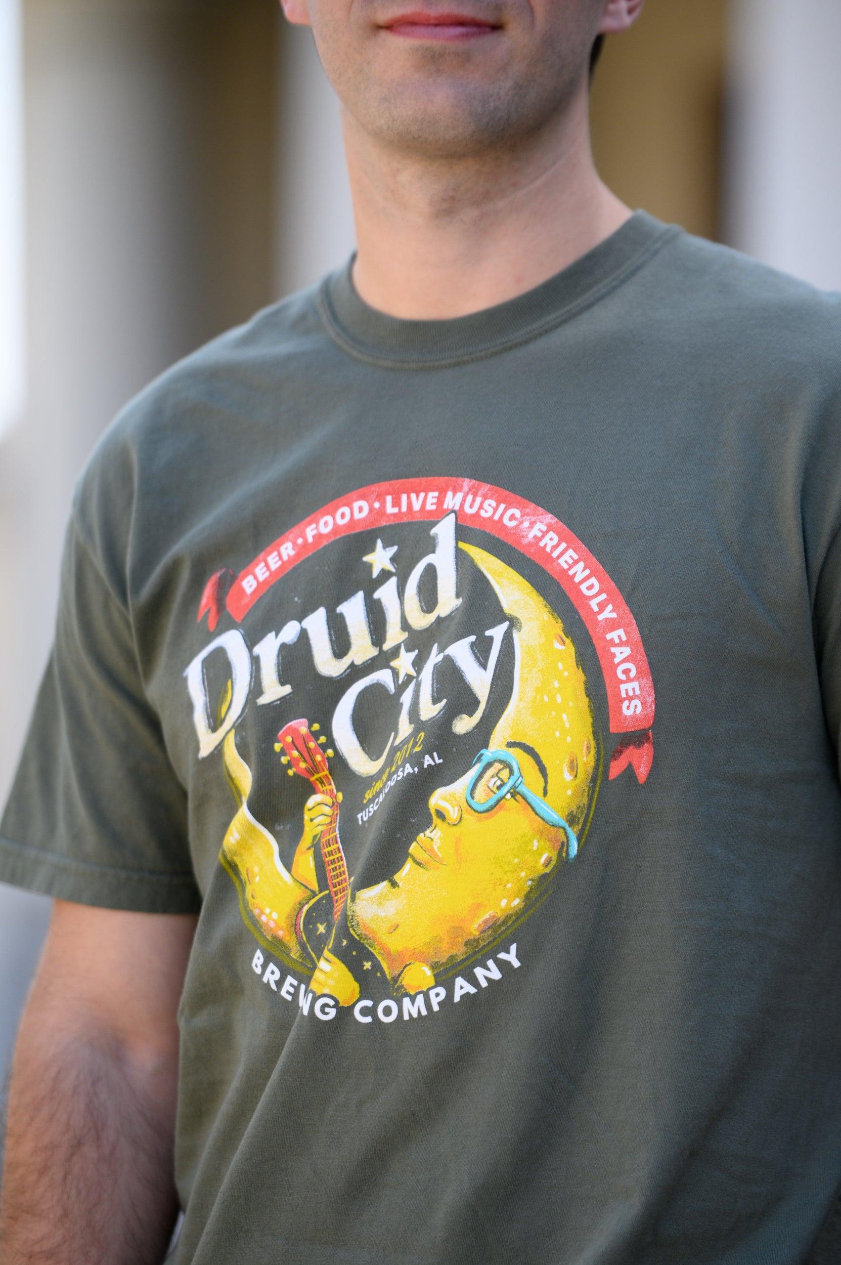 Druid City Brewing Company Olive Green Tee with text "Druid City" above a Crescent Moon wearing glasses and playing a guitar