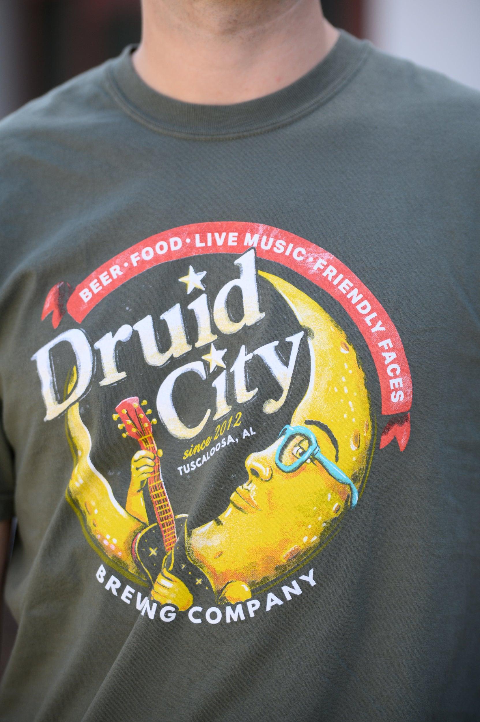 Druid City Brewing Company Olive Green Tee with text "Druid City" above a Crescent Moon wearing glasses and playing a guitar