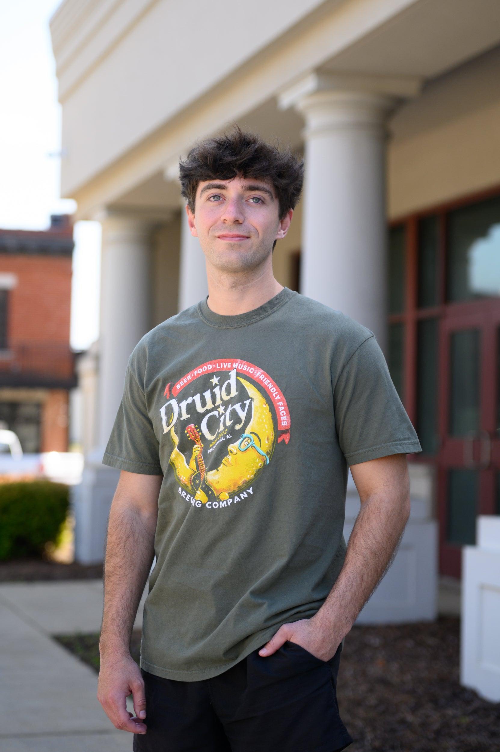 Druid City Brewing Company Olive Green Tee with text "Druid City" above a Crescent Moon wearing glasses and playing a guitar