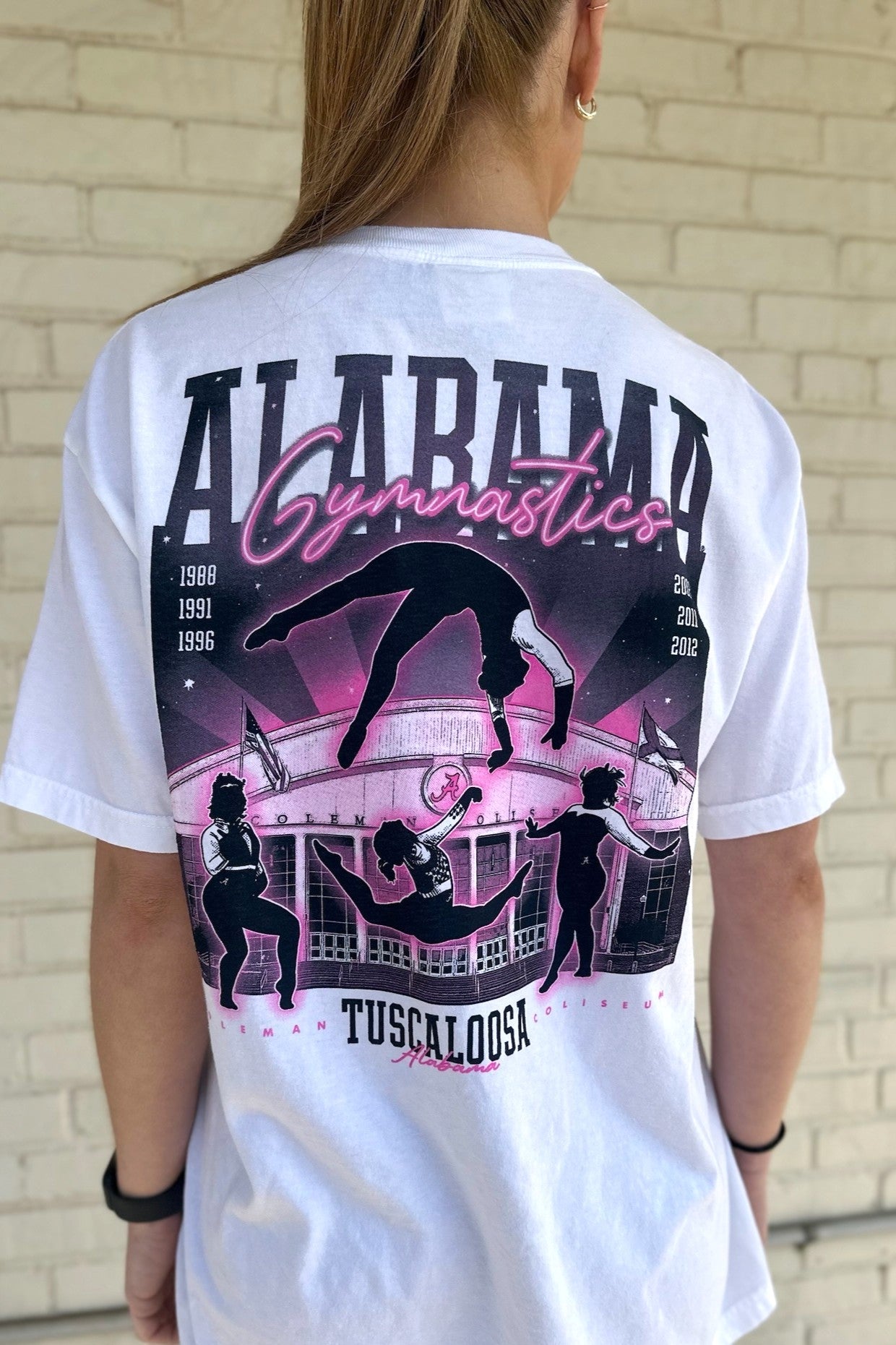 University of Alabama White Ladies' Gymnastics T-Shirt with a hand-drawn design featuring Coleman Coliseum and the years of the Crimson Tide's national titles.