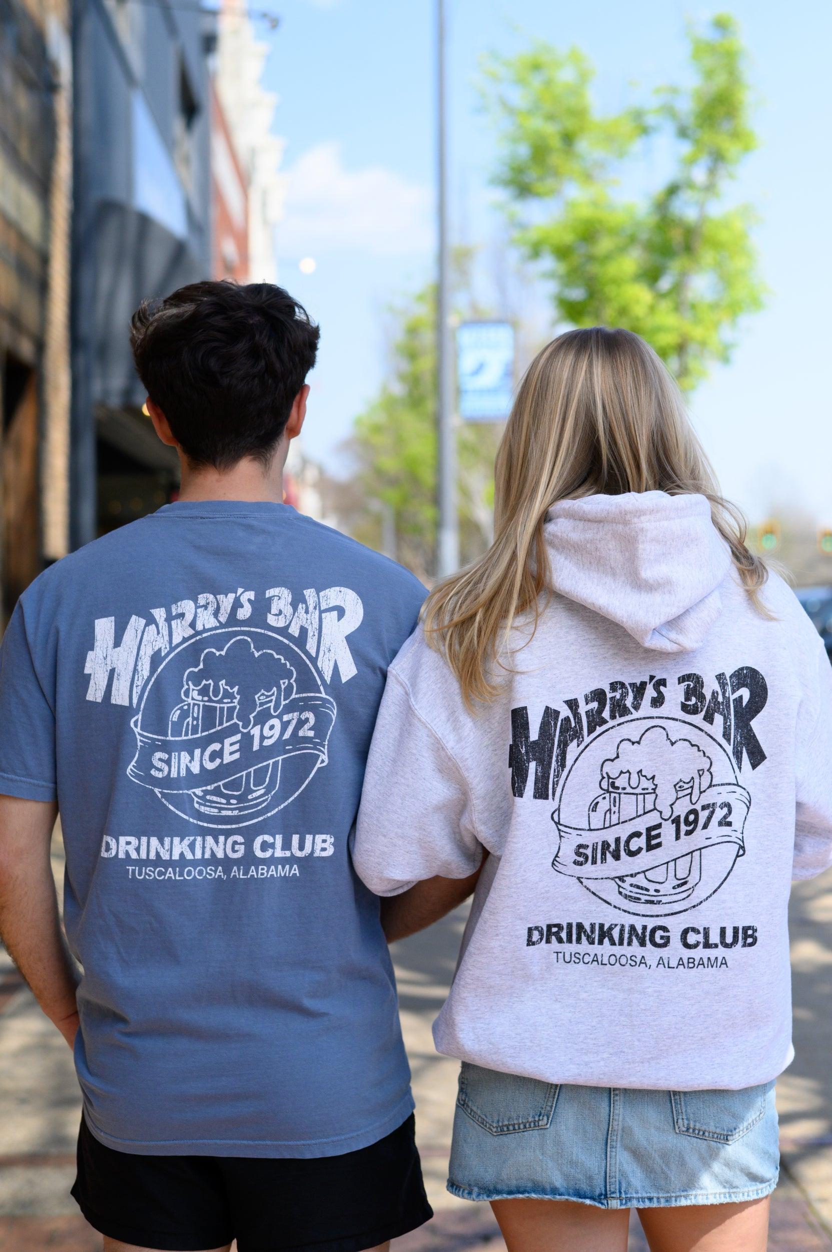 Two people wearing shirts with 'Harry's Bar Drinking Club' design on a street.