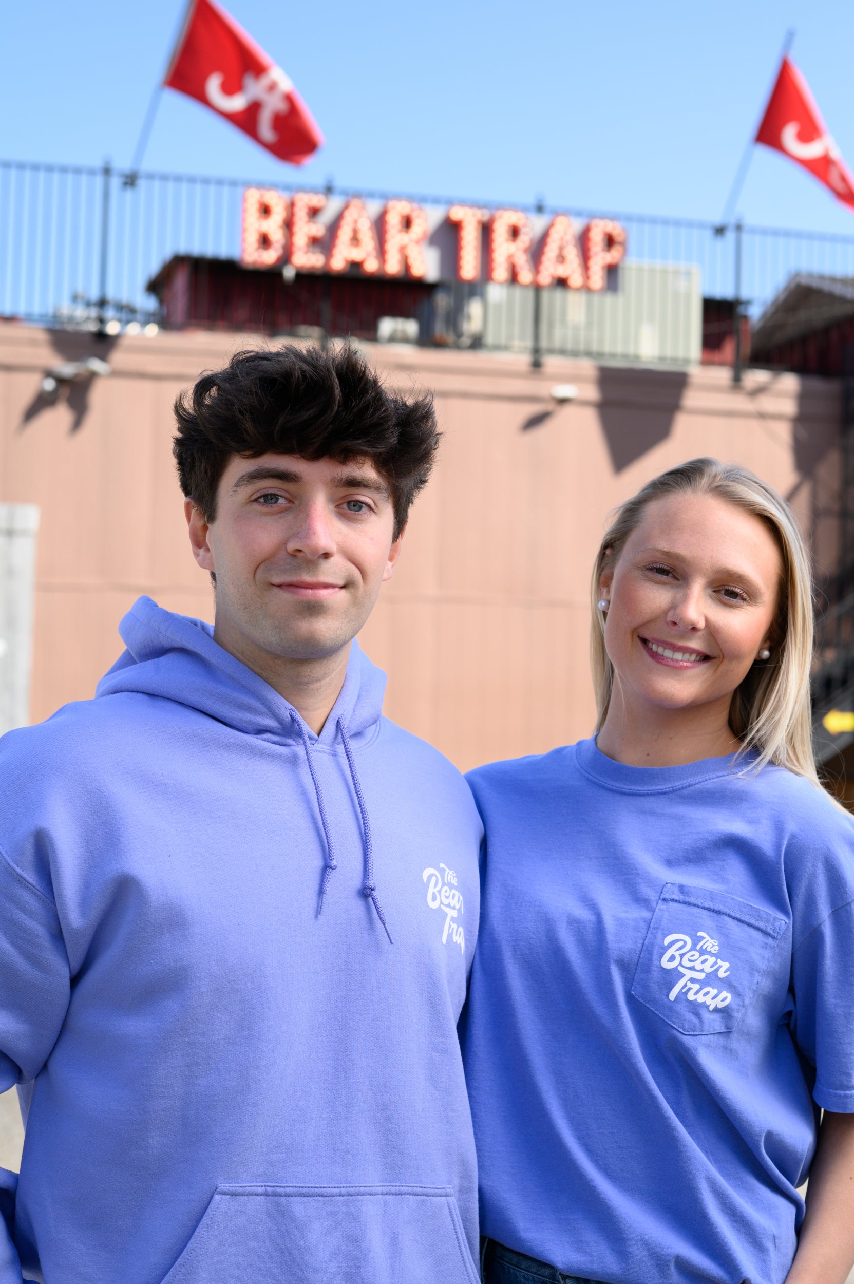 Two people wearing blue hoodies and t-shirts with 'Bear Trap' branding in front of a the Bear Trap Building
