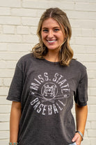 Woman wearing a dark gray t-shirt with 'Mississippi State Baseball' text against a white brick wall.