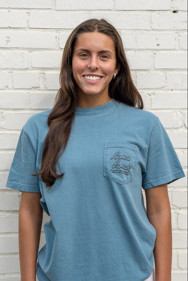 Woman wearing a blue t-shirt with a pocket design against a white brick wall.