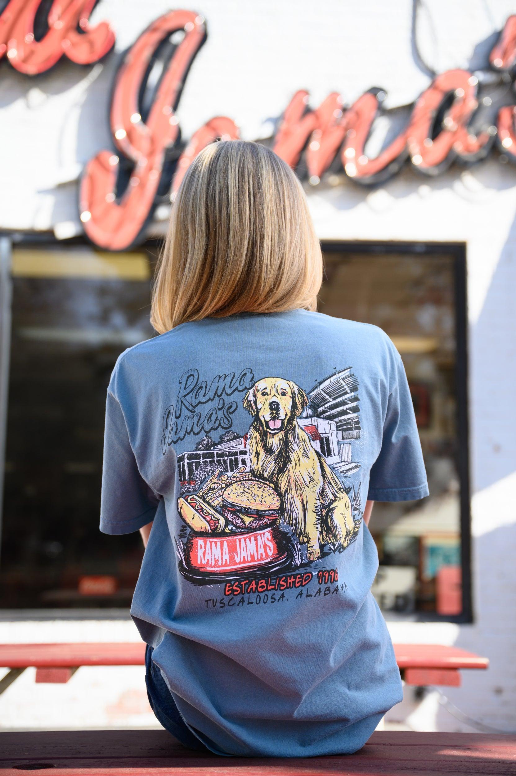 Rama Jama’s Dusty Blue shirt with an illustration of a golden retriever sitting at the picnic tables outside of the building with a cheeseburger and a hotdog