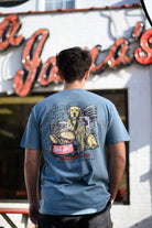 Rama Jama’s Dusty Blue shirt with an illustration of a golden retriever sitting at the picnic tables outside of the building with a cheeseburger and a hotdog
