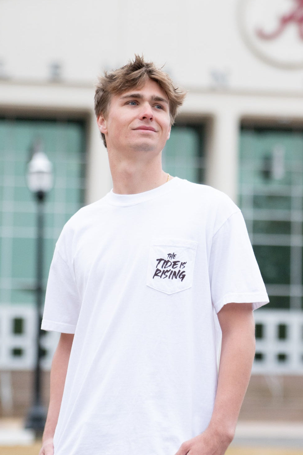 University of Alabama White T-shirt with a Script A brand folding chair that has a red flannel hanging over the back and a play planning board leaning against it. Below, Black text reading “The Tide is Rising”