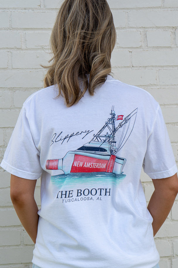 Person wearing a white t-shirt with a graphic design of a boat and text, standing against a light brick wall.