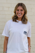 Woman wearing a white t-shirt with a logo, standing against a light brick wall.