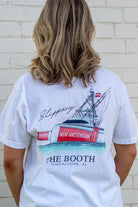 Person wearing a white t-shirt with a graphic design of a ship and text on the back, standing against a white brick wall.
