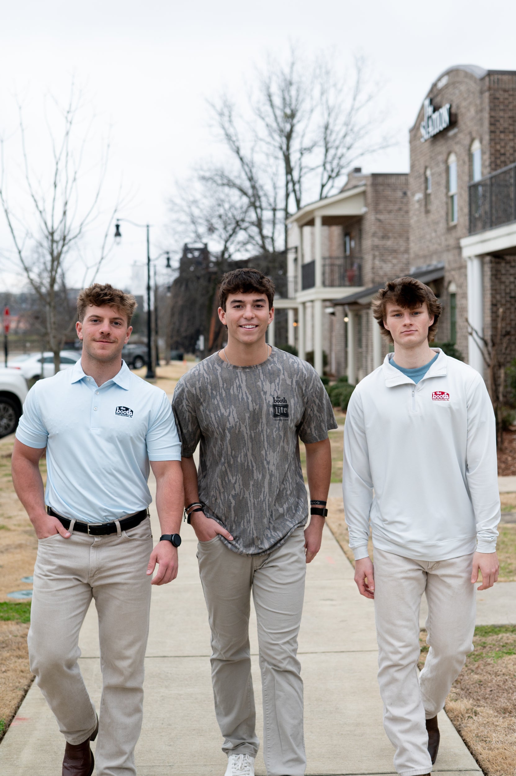 Three young men standing on a sidewalk in front of a building wearing "The Booth" branded products.