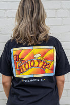 Person wearing a black t-shirt with colorful graphic design on the back, standing against a white brick wall.