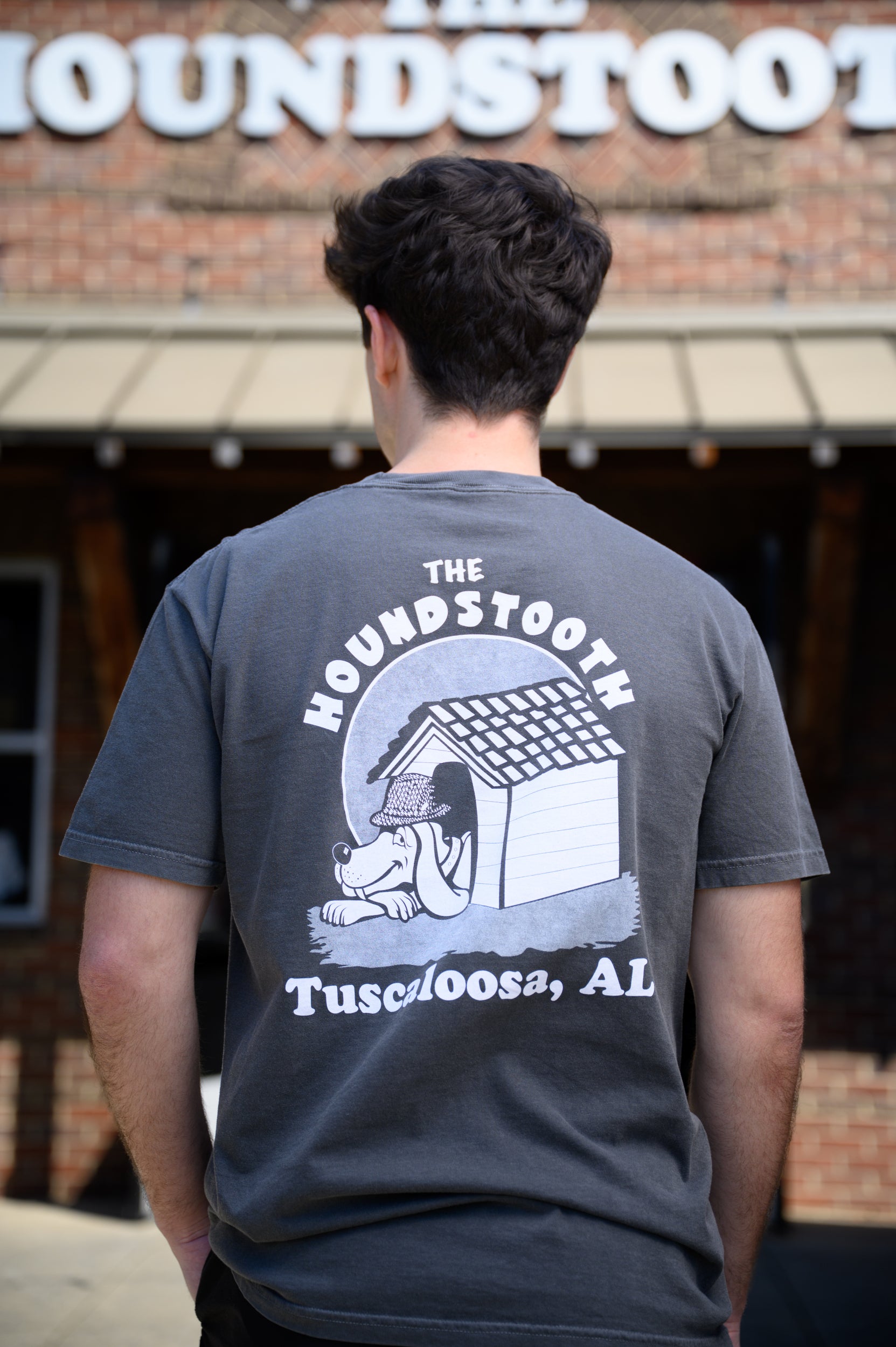 The Houndstooth Dark Grey T-Shirt with Black text "The Houndstooth" above an image of a Beagle laying inside of a doghouse