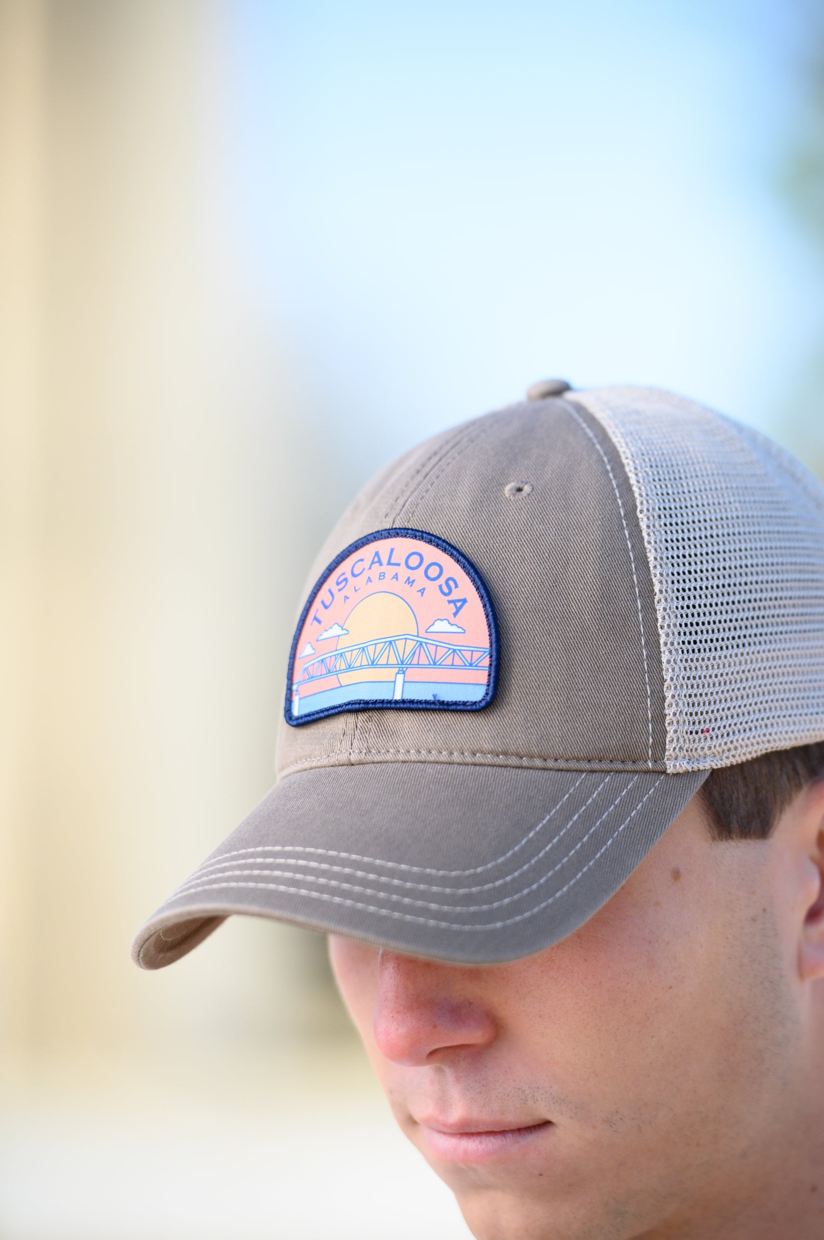 Tuscaloosa Beige Trucker Hat with a patch featuring Navy text "Tuscaloosa Alabama" a drawing of the sun setting on the iconic Tuscaloosa Trestle