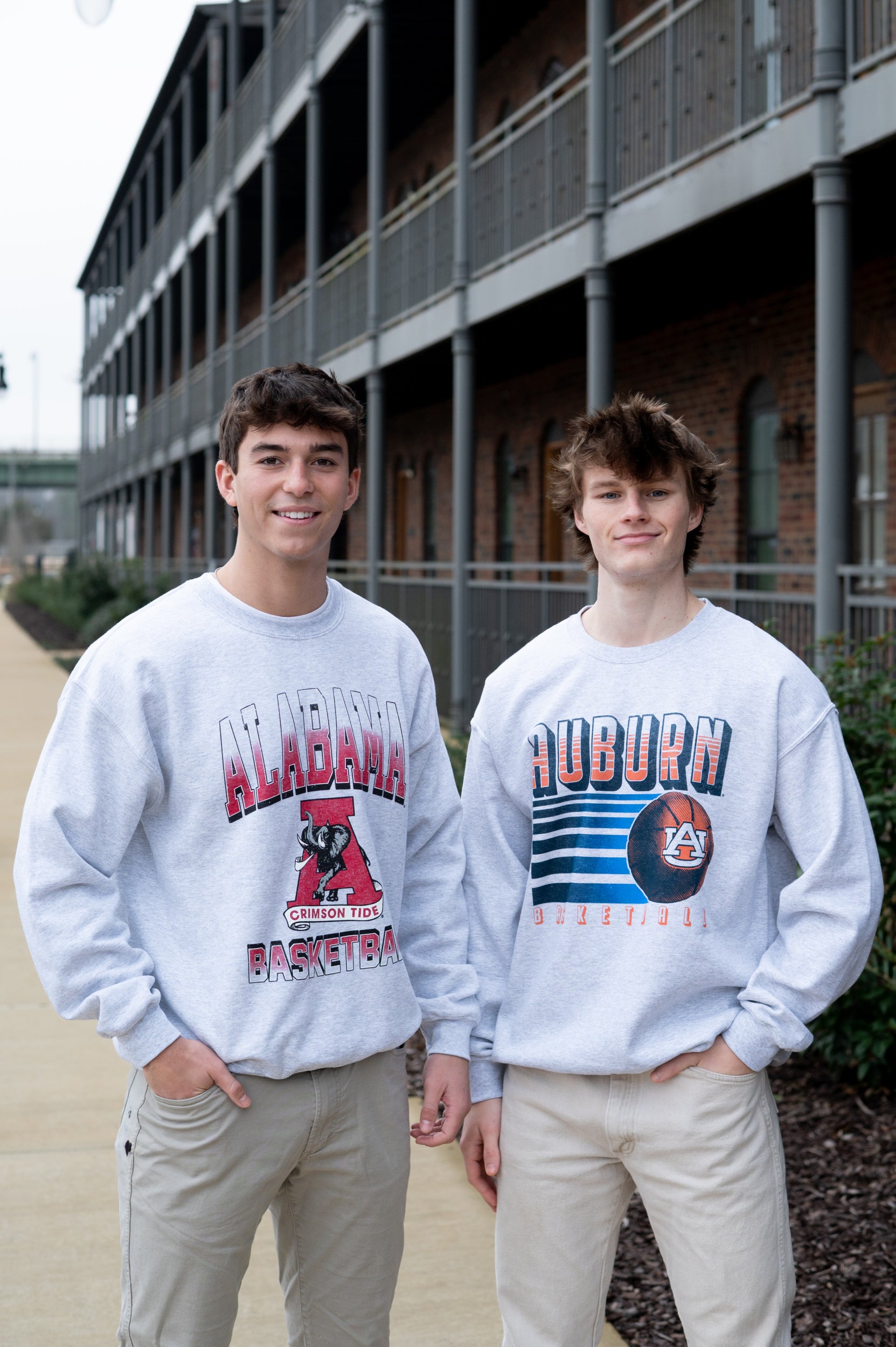 Two individuals wearing gray sweatshirts with 'Alabama' and 'Auburn' logos in front of a building.