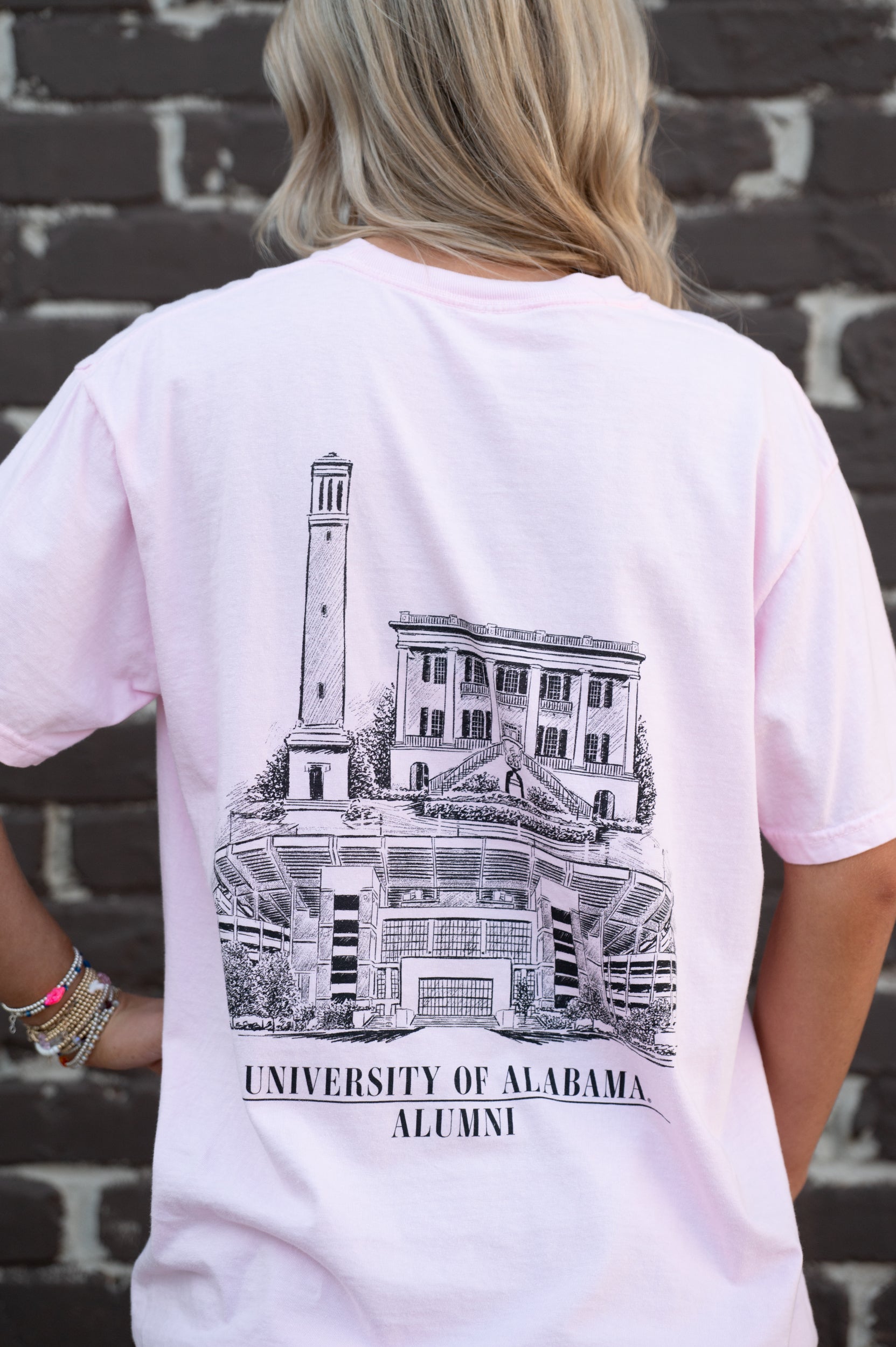 University of Alabama Alabama Alumni pink Comfort Colors T-Shirt with a graphic of Bryant-Denny Stadium, the President's Mansion, and Denny Chimes, and the text 'UNIVERSITY OF ALABAMA ALUMNI'. 