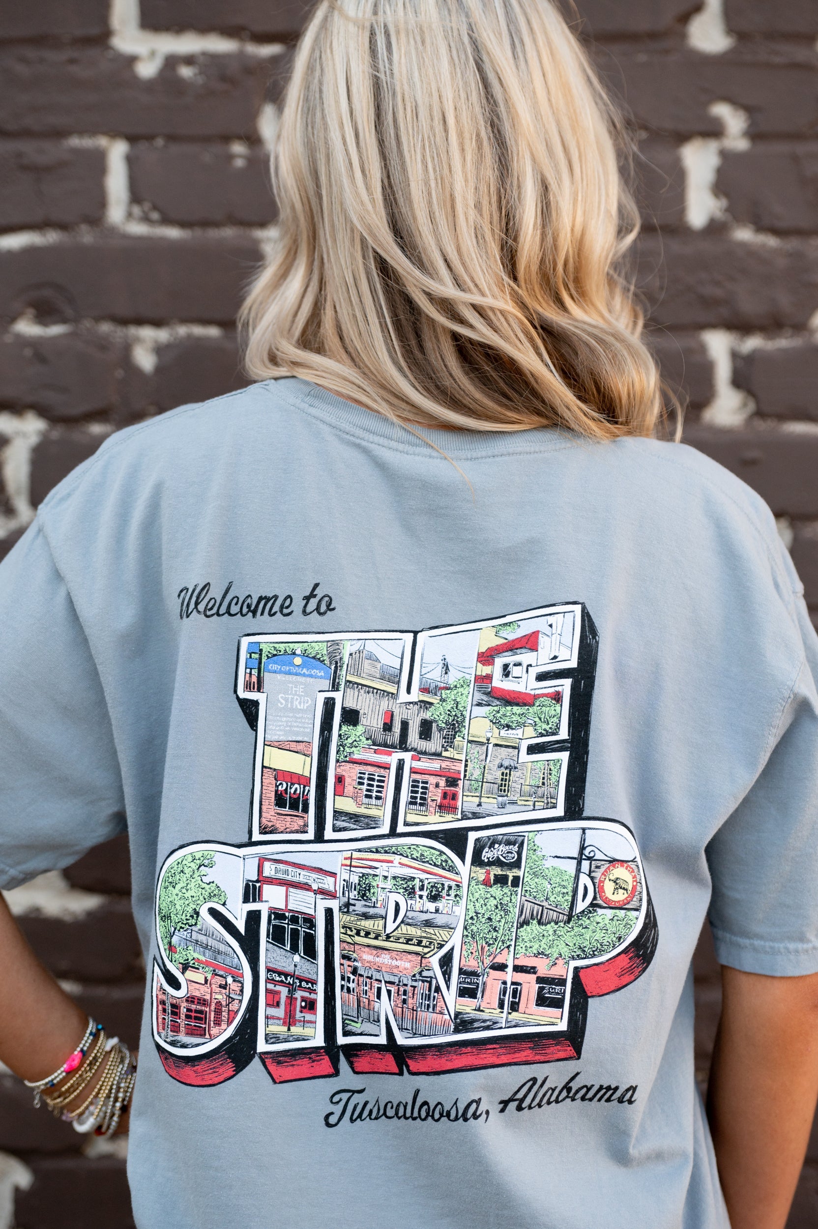 University of Alabama Grey The Strip T-Shirt with postcard style text "Welcome to THE STRIP" with "The Strip" filled with images of iconic buildings in that location